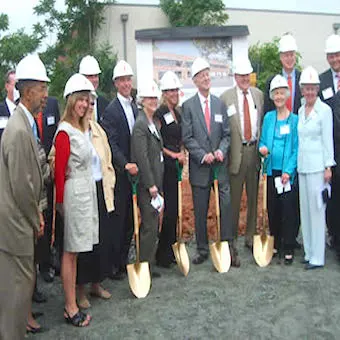 A group of people wearing hard hats and business attire stand together outdoors, smiling and holding gold shovels for a groundbreaking ceremony. A building plan is displayed behind them.