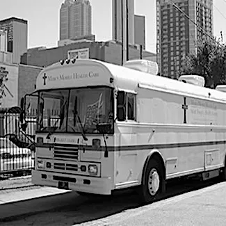 A white mobile health care bus with a cross symbol is parked on a city street, surrounded by tall buildings and a fence, in a black-and-white photo.