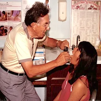 An older man uses a medical instrument to examine a young girls throat in a clinic, with posters and medical supplies visible in the background.