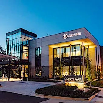 A modern medical building labeled Mercy Care and Mercy Park is lit up at dusk. The structure features large glass windows, a well-lit entrance, and landscaped grounds in front.