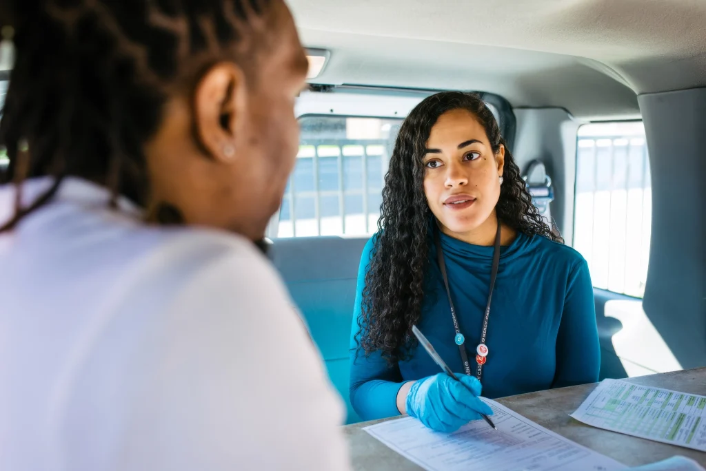 A woman wearing blue gloves and a lanyard, likely one of the clinical volunteers, speaks to a man while holding a pen and a form inside a vehicle, suggesting an interview or assistance situation.