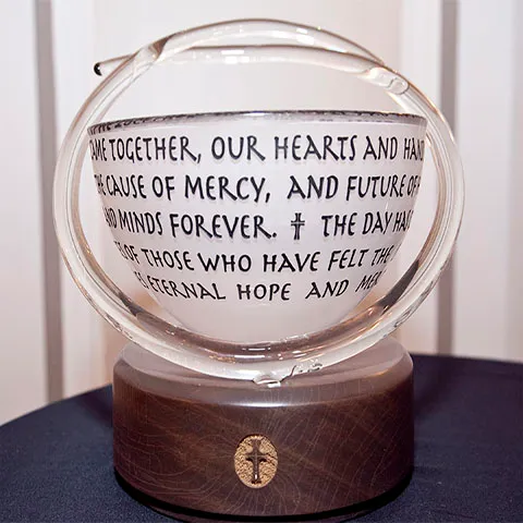A clear glass globe with handwritten text inside sits on a wooden base featuring a small cross symbol. The text inside reads a message about unity, mercy, and hope. The globe is displayed on a dark cloth.