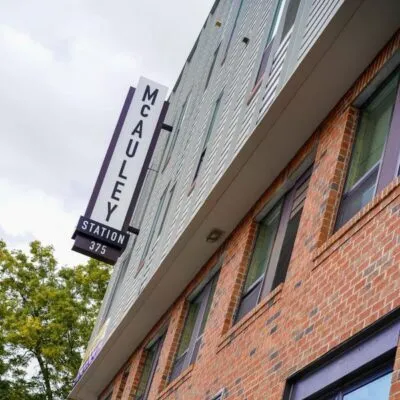 A multi-story brick and siding building with a vertical sign reading McAuley Station 375 mounted above the entrance, viewed from a low angle against a cloudy sky.