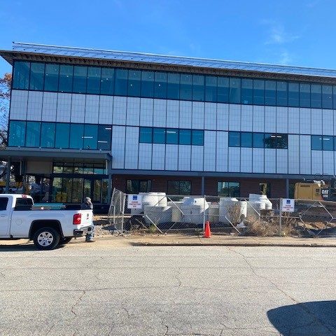 A modern three-story building under construction, with large glass windows and white paneling. A white truck and construction equipment are in front, and a fence surrounds the site. A person walks near the entrance.