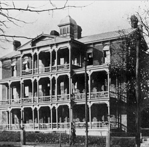 Black-and-white photo of a large, historic three-story building with multiple balconies, ornate railings, and a central cupola. Leafless tree branches frame the image, and a fence is visible in front.