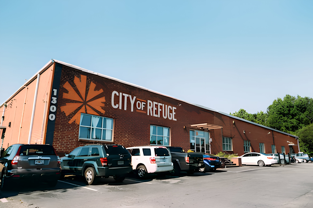 A brick building with CITY OF REFUGE painted on the side, next to an orange sunburst logo. Several cars are parked in front, and trees are visible in the background under a clear blue sky.