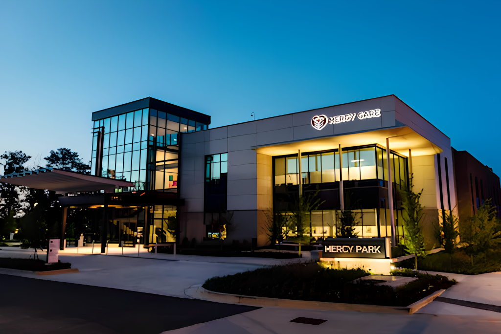A modern healthcare building labeled “Mercy Care” and “Mercy Park” is lit up at dusk, with large glass windows and landscaped surroundings. The sky is clear, and lights illuminate the entrance and exterior.