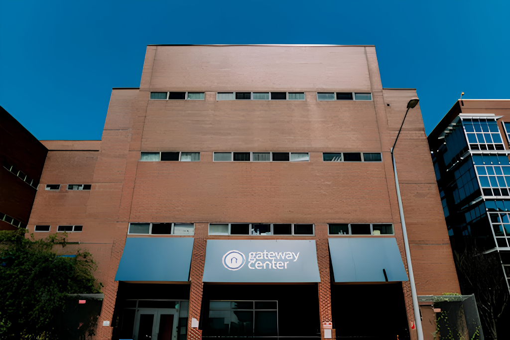 A large brick building with multiple windows and blue awnings. A sign above the entrance reads gateway center. The sky is clear and blue, and a lamppost stands on the right side of the image.