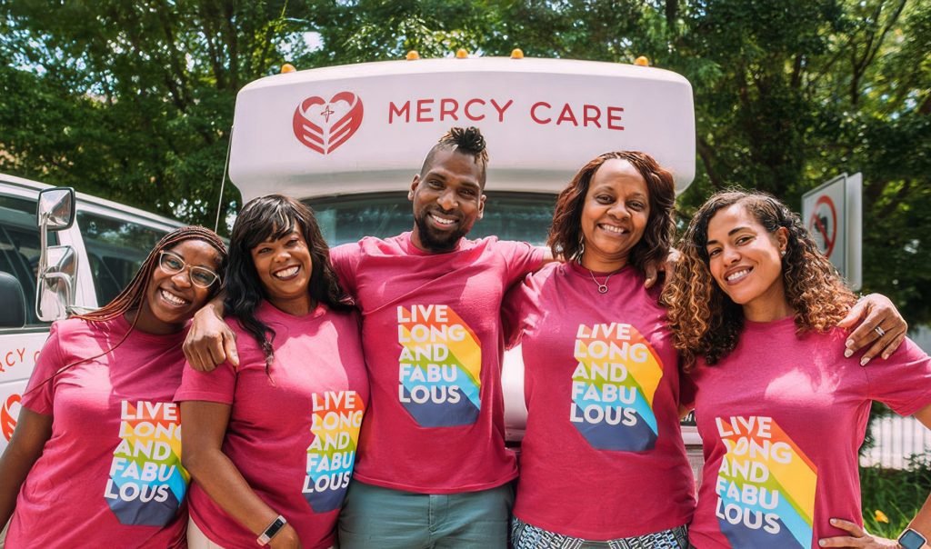 Five smiling people stand arm-in-arm in front of a Mercy Care van, all wearing pink shirts with a rainbow design and the words Live Long and Fabulous. Trees and greenery are visible in the background.
