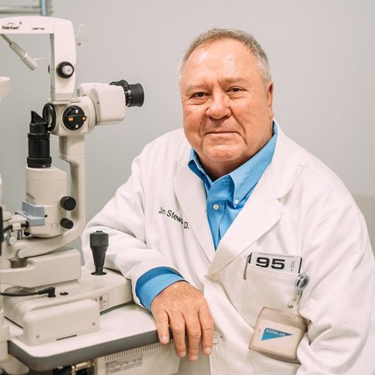 An older male doctor in a white lab coat sits beside ophthalmology equipment, smiling at the camera in a medical examination room.