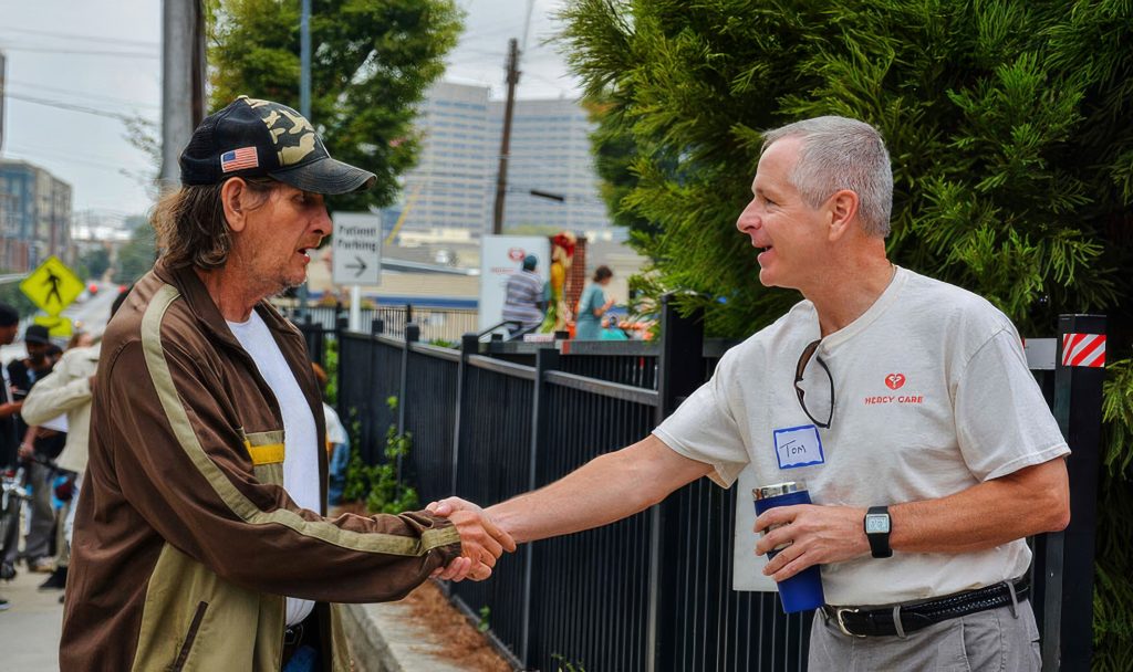 A man in a camouflage hat and jacket shakes hands with another man in a Mercy Care shirt holding a cup. They are outdoors near a fence, with greenery and city buildings in the background.