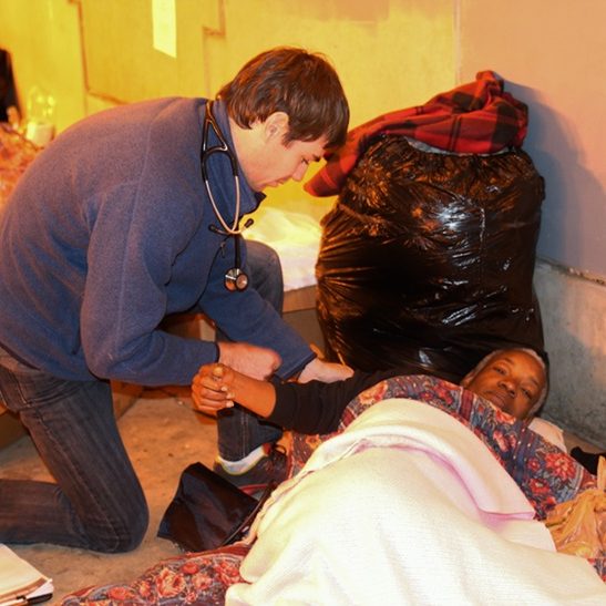 A healthcare worker kneels beside a patient lying on a makeshift bed, checking their blood pressure in an indoor shelter setting with basic bedding and personal belongings nearby.
