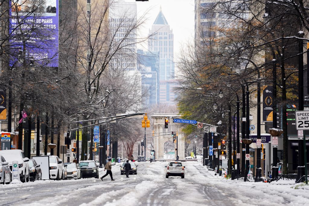 A snow-covered city street with cars parked along the sides, people walking across the road in cold weather, and tall buildings in the background. Trees and traffic lights line the street on a wintry day.