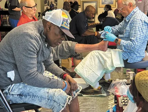 Several people sit in chairs while Mercy Care volunteers provide foot washing and tend to their feet. The setting appears to be a community service event for Atlanta homeless, with supplies and equipment visible around them.