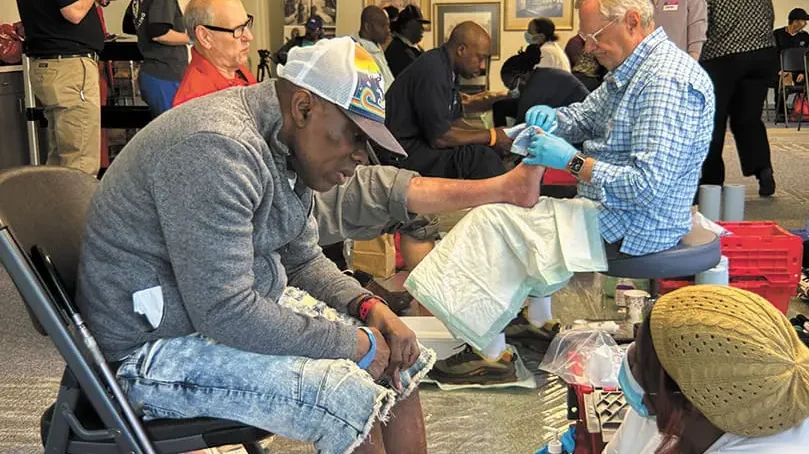 Several people sit in chairs while Mercy Care volunteers provide foot washing and tend to their feet. The setting appears to be a community service event for Atlanta homeless, with supplies and equipment visible around them.