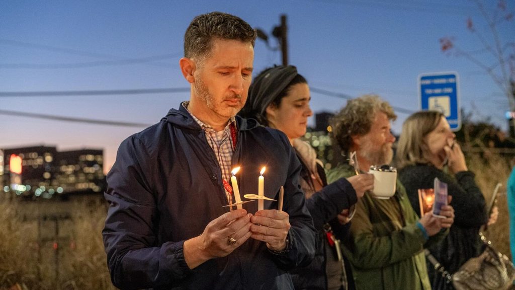 A group of people stand outdoors at dusk holding lit candles during a Candlelight Memorial, appearing somber and reflective. Buildings and a blue sign honoring Robert L. Mason Jr. are visible in the background.