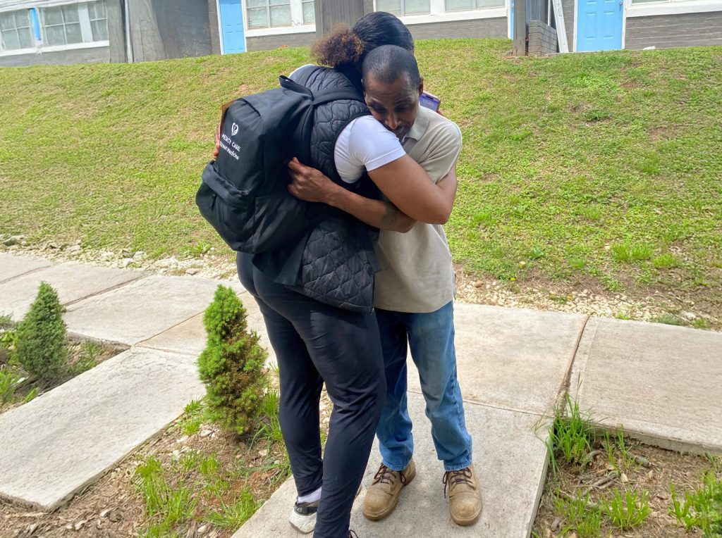 Two people stand on a sidewalk outside an apartment building near Mercy Care Atlanta, hugging each other tightly. One person wears a black backpack and jacket; the other wears a light shirt and jeans. Grass and small shrubs are visible nearby.