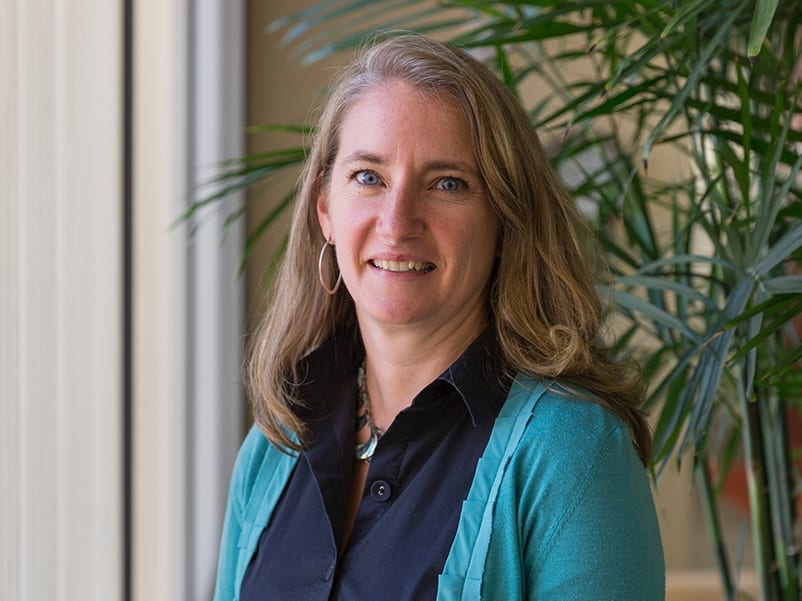 Kathryn Lawler, recognized in Atlanta 500 for her impact in Education & Healthcare, stands indoors near a window and green plants, smiling at the camera with long light brown hair and a teal cardigan over a black shirt.